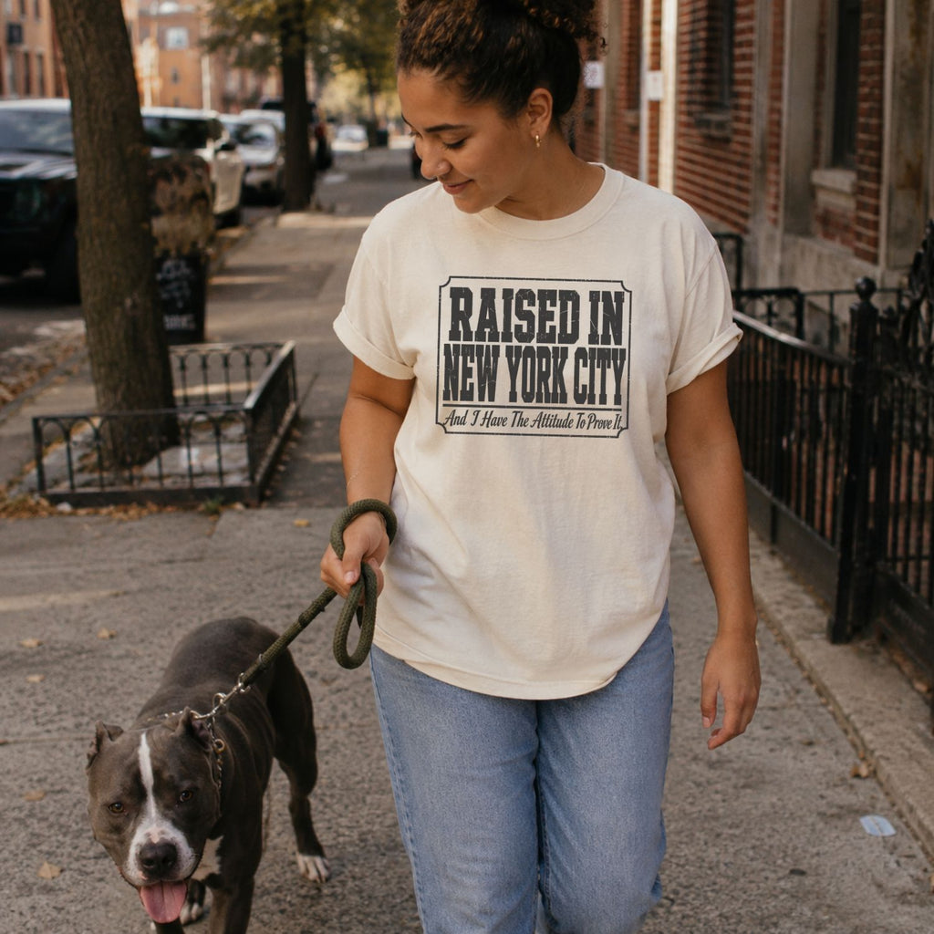 Woman walking a dog on a city street wearing a 'Raised in New York City' t-shirt.