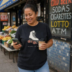 Woman holding a phone and coffee cup in front of a store with a sign for lottery and ATM wearing a black Comfort Colors t-shirt featuring a vintage roller skate graphic with “Roll With It • NYC • 1988” text.