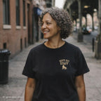 Woman wearing a black Comfort Colors t-shirt with the words "Skate Key Staff" and roller skate graphic on the left chest, standing on a city street with brick buildings in the background. 