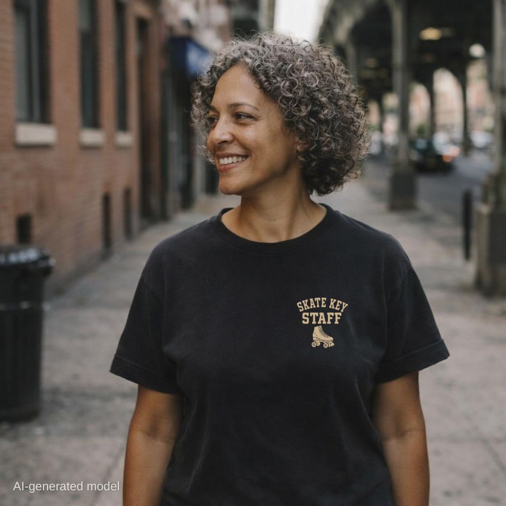 Woman wearing a black Comfort Colors t-shirt with the words "Skate Key Staff" and roller skate graphic on the left chest, standing on a city street with brick buildings in the background. 