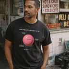 Man wearing a black Comfort Colors T-shirt with NYC Spaldeen specimen front print, while standing in front of a deli
