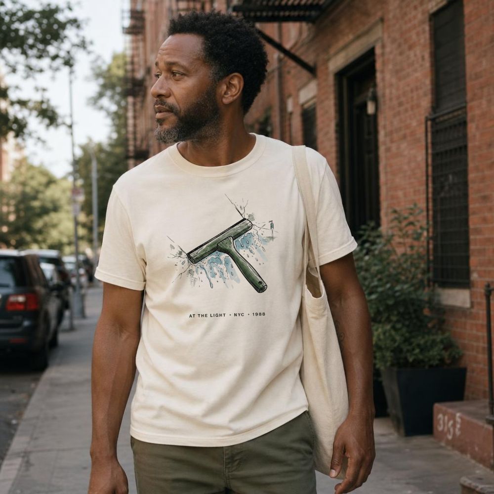 Man wearing an Ivory Comfort Colors t-shirt with graphic design of a squeegee and text on a city street.