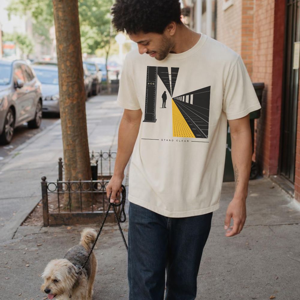 Man walking a dog on a city street wearing an Ivory Comfort Colors t-shirt with a graphic design of a person on train tracks.