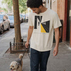 Man walking a dog on a city street wearing an Ivory Comfort Colors t-shirt with a graphic design of a person on train tracks.
