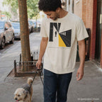 Man walking a dog on a city street wearing an Ivory Comfort Colors t-shirt with a graphic design of a person on train tracks.