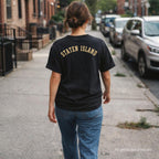 Woman wearing a black Comfort Colors t-shirt with 'Staten Island' text, walking on a sidewalk.