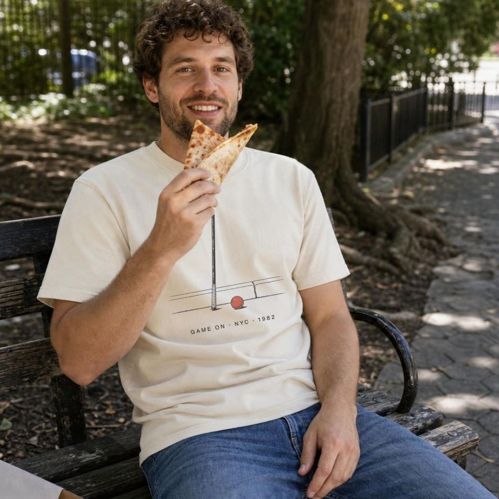 Man sitting on a bench outdoors, holding a slice of pizza and wearing a Ivory Comfort Colors t-shirt featuring a minimalist stickball bat and ball graphic with “Game On • NYC • 1982” text.