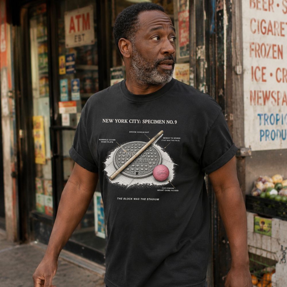 Man wearing a black Comfort Colors t-shirt with a stickball graphic design, standing in front of a store.
