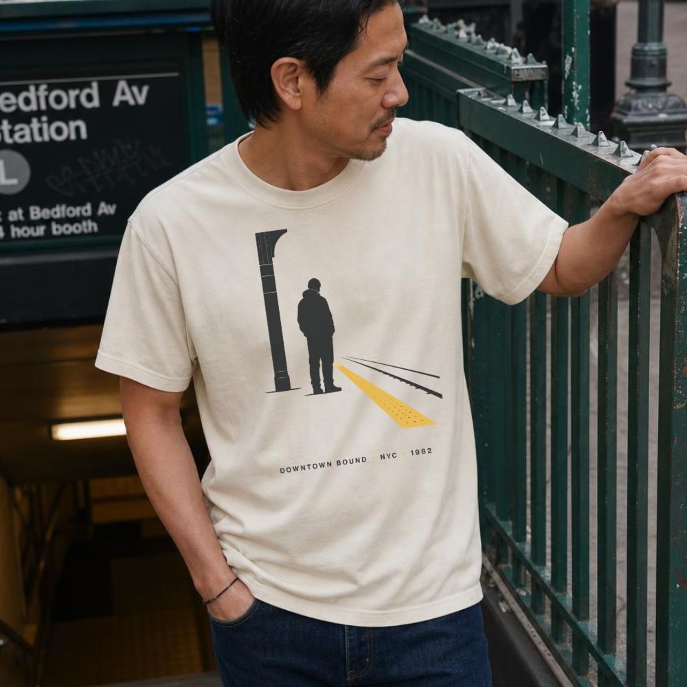 Man wearing an Ivory Comfort Colors t-shirt with a graphic design of a person on a train platform, standing near at a subway entrance.