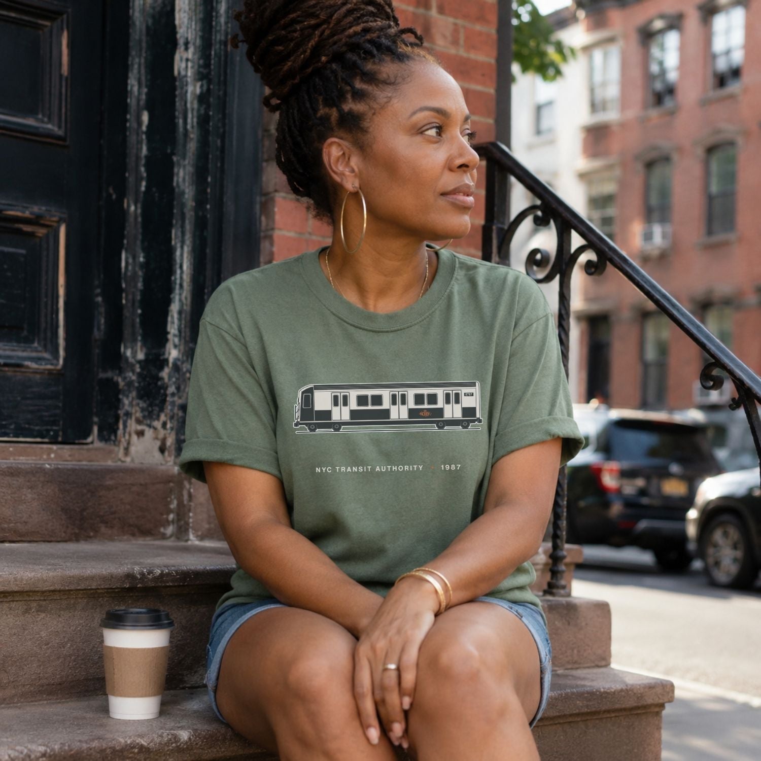 Woman sitting on a stoop wearing a Moss green Comfort Colors t-shirt featuring a minimalist New York City subway car graphic with “NYC Transit Authority • 1987” text, holding a coffee cup.
