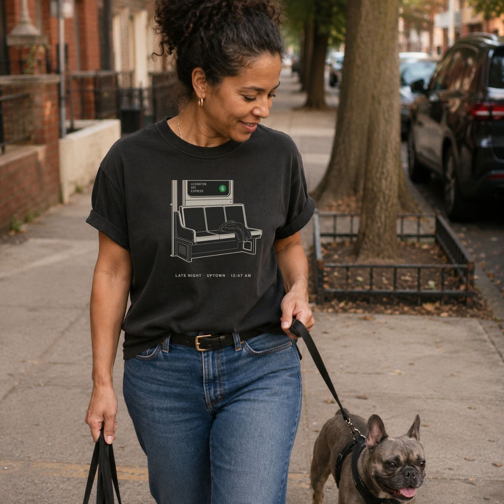 Woman walking a dog on a leash in an urban setting, wearing a Black t-shirt with a graphic design of a train seat and text.