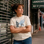 Woman wearing an Ivory Comfort Colors t-shirt featuring a vintage NYC fire hydrant spraying water with “Summer Days • NYC • 1981” text, placed on a dark surface with a denim jacket and a map, standing in front of a laundromat.