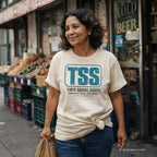 A woman wearing an ivory Comfort Colors graphic T-shirt with a blue 'TSS Times Square Stores' logo, standing on a city street in front of a store entrance.