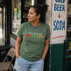 Woman wearing a Moss green Comfort Colors t-shirt featuring a minimalist Bronx subway line graphic with The Bronx text and colorful NYC train icons, standing in front of a store sign.