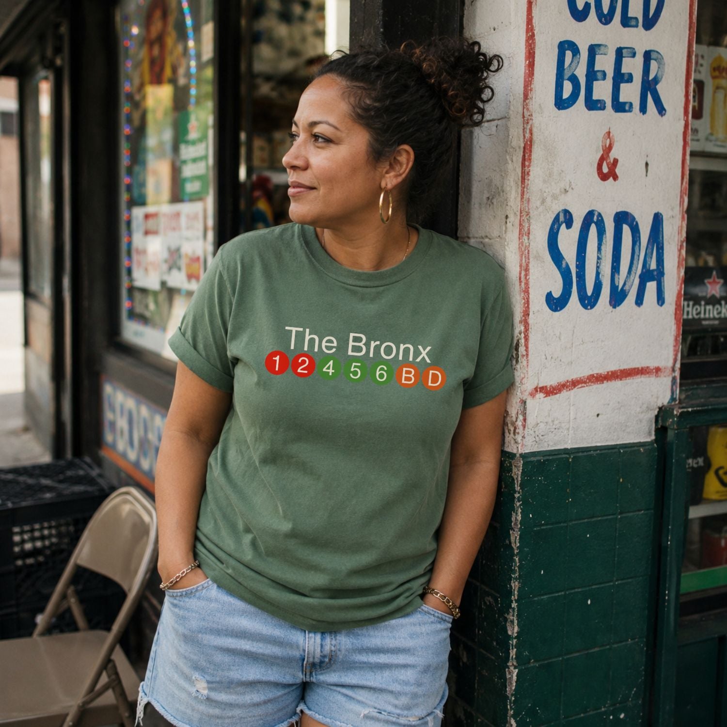 Woman wearing a Moss green Comfort Colors t-shirt featuring a minimalist Bronx subway line graphic with The Bronx text and colorful NYC train icons, standing in front of a store sign.