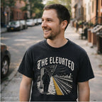 Man wearing a Black Comfort Colors t-shirt with gothic lettering reading "The Elevated" arched over a detailed illustration of a Bronx elevated train platform in winter, single figure in a puffer coat facing empty tracks with a yellow safety stripe. while standing on the sidewalk on a NYC street. 