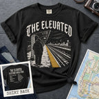 Black Comfort Colors t-shirt with gothic lettering reading "The Elevated" arched over a detailed illustration of a Bronx elevated train platform in winter, single figure in a puffer coat facing empty tracks with a yellow safety stripe. Back print lists all 12 stops on the 4 train from 161st St/Yankee Stadium to Woodlawn with "The Bronx. IRT 4 Train. Since 1917" at the bottom.