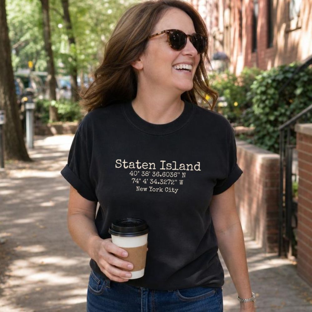 Woman wearing a black Comfort Colors t-shirt with Staten Island text and coordinates, holding a coffee cup outdoors.