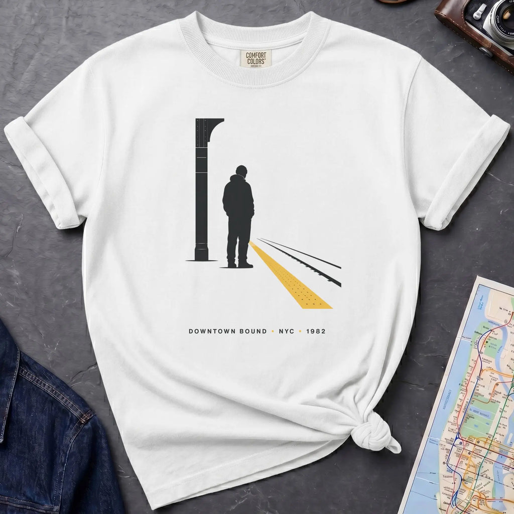 White t-shirt with a graphic design of a person on a train platform, placed on a dark surface with a map and denim jacket.