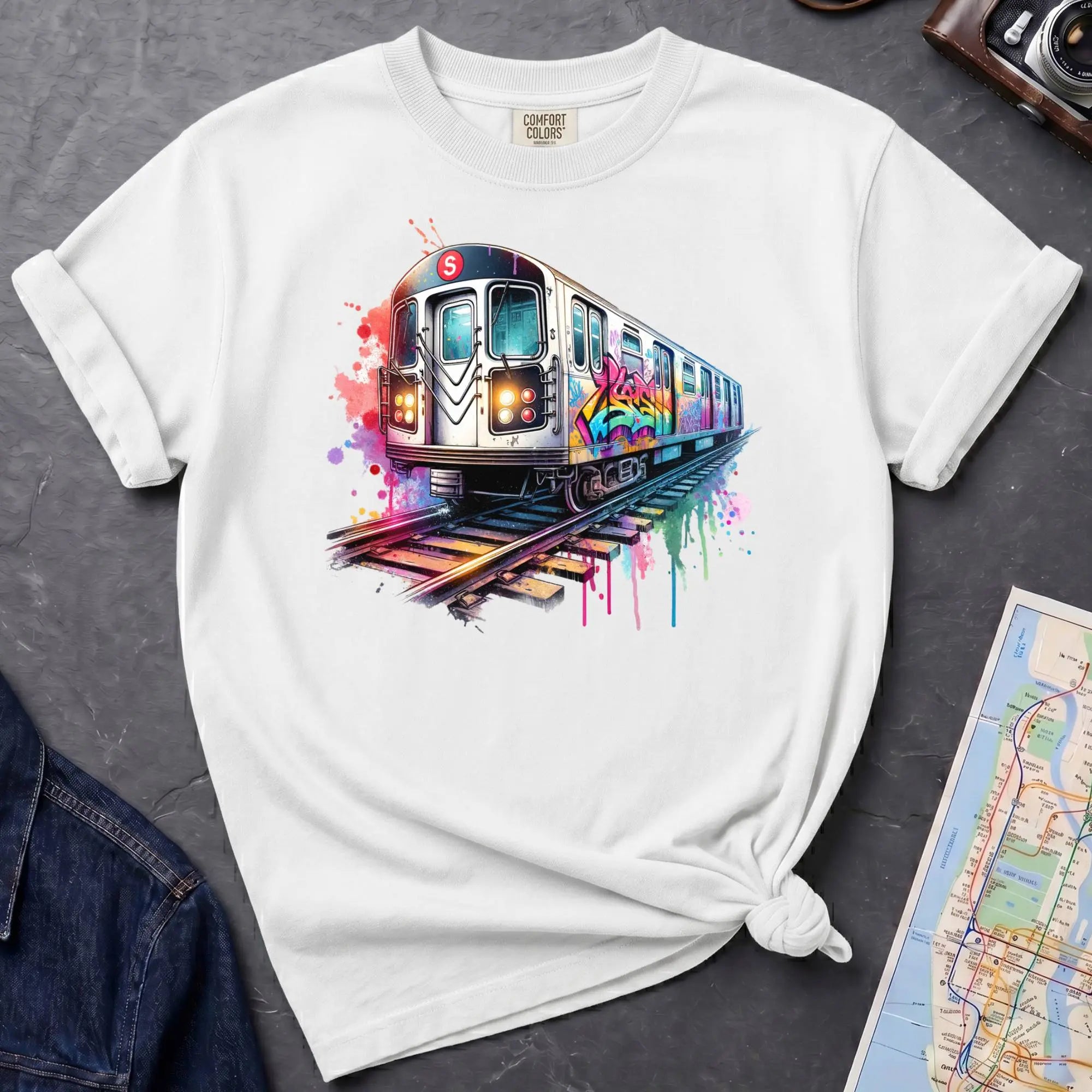 White Comfort Colors t-shirt featuring a colorful NYC subway train graphic with graffiti-style paint details and bright street art accents.