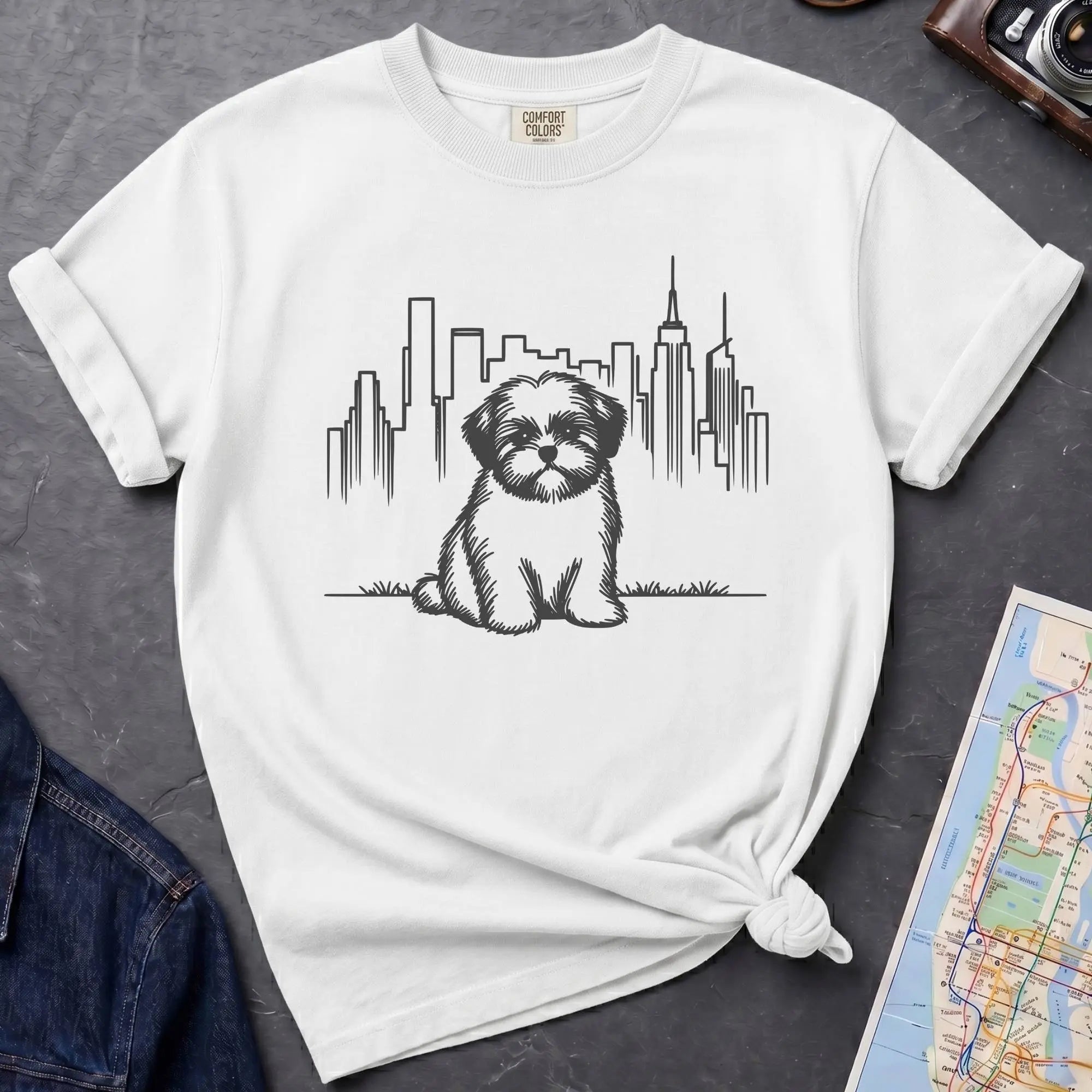 White t-shirt with a Shih Tzu dog and cityscape design on a dark surface with a map and camera.