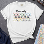White t-shirt with 'Brooklyn' and subway line numbers printed on it, placed on a dark surface with a map and denim jacket.
