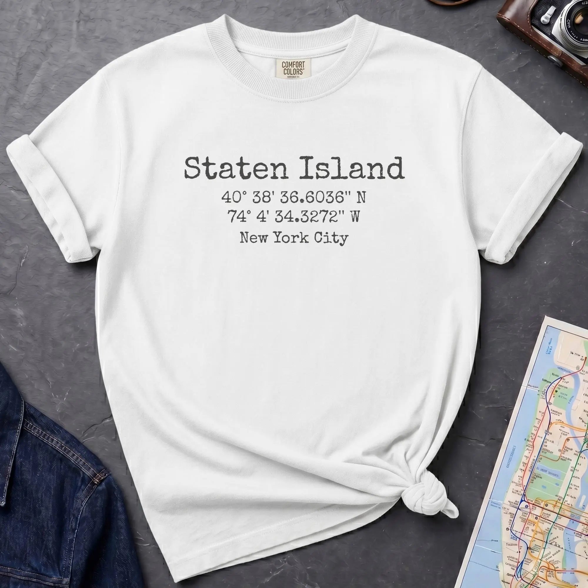 White t-shirt with Staten Island text and coordinates on a dark surface with denim jacket and map.
