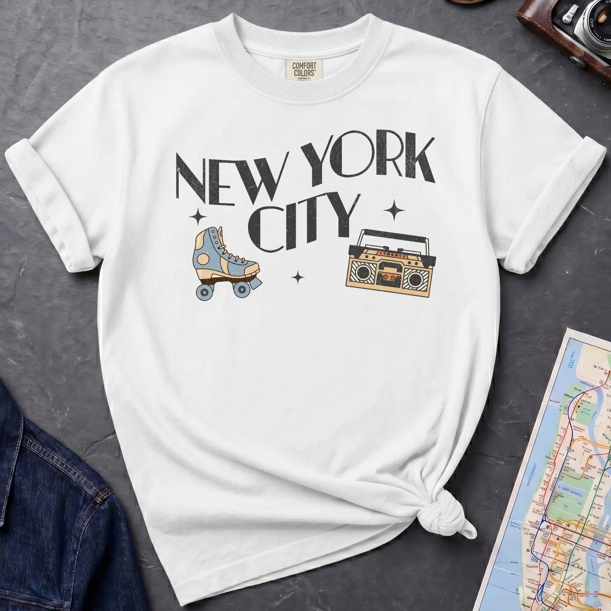 White t-shirt with 'New York City' text and a roller skate and boom box on a dark surface with a map and camera.