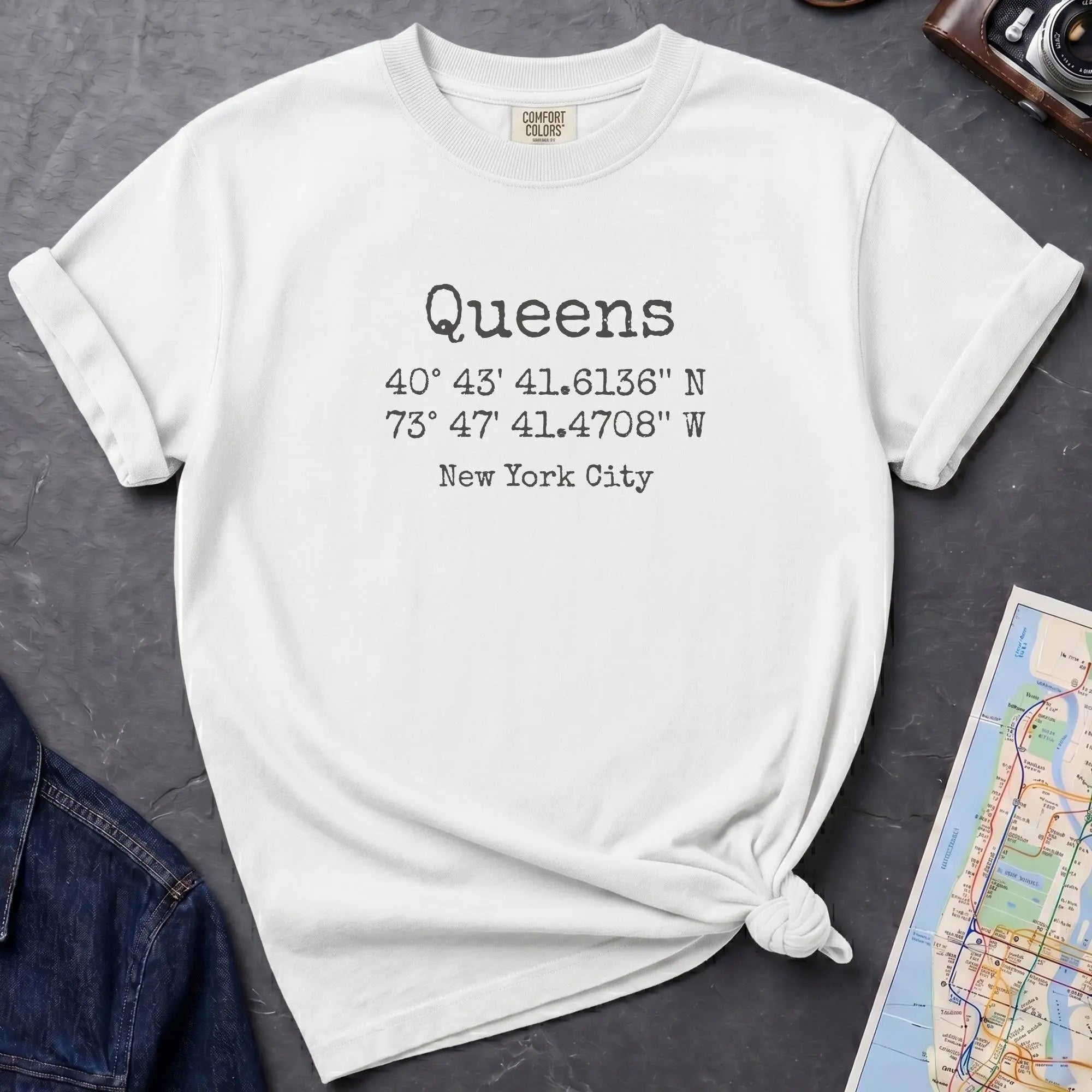 White t-shirt with 'Queens' text and coordinates, placed on a dark surface with a map and denim jacket.