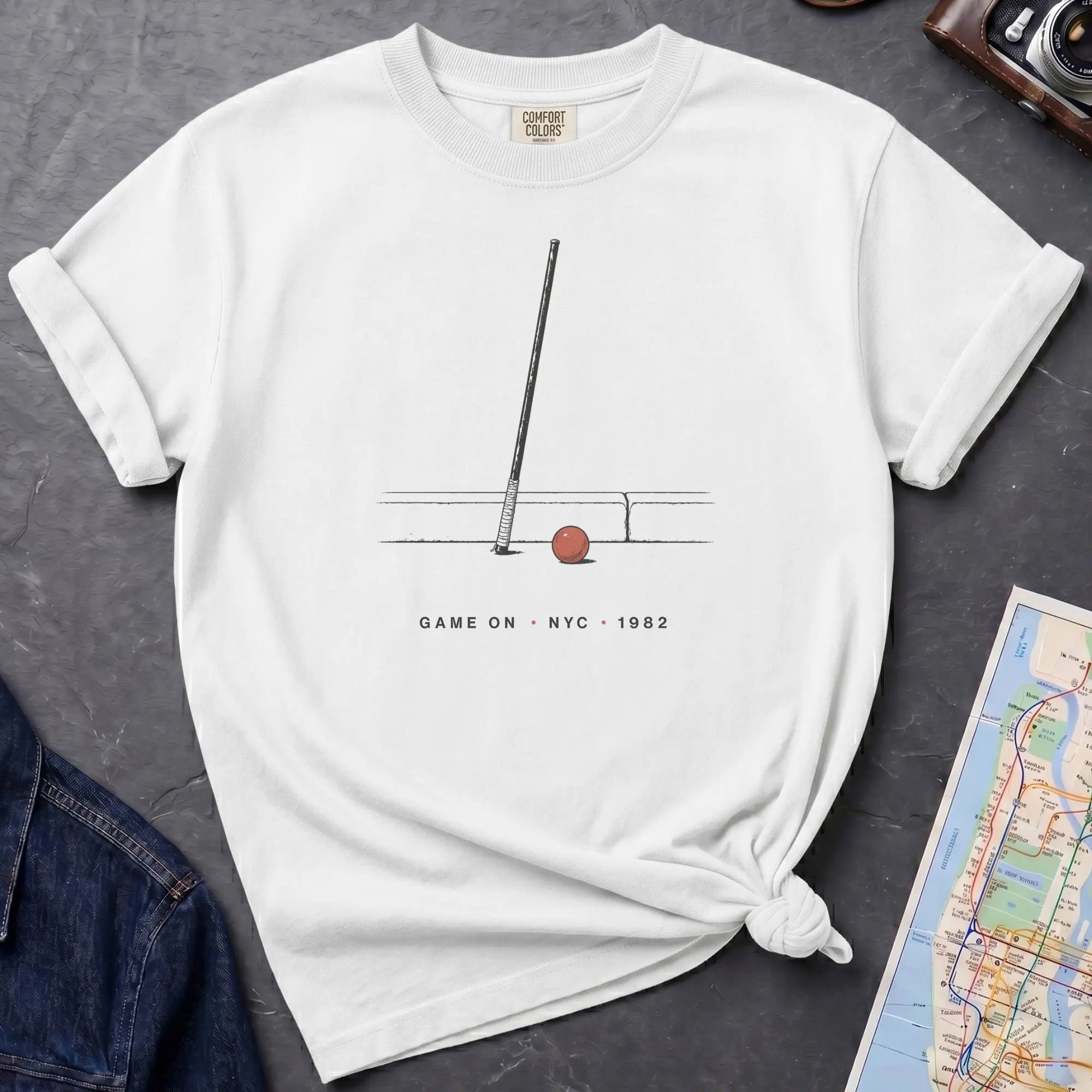 White Comfort Colors t-shirt featuring a minimalist stickball bat and ball graphic with “Game On • NYC • 1982” text.