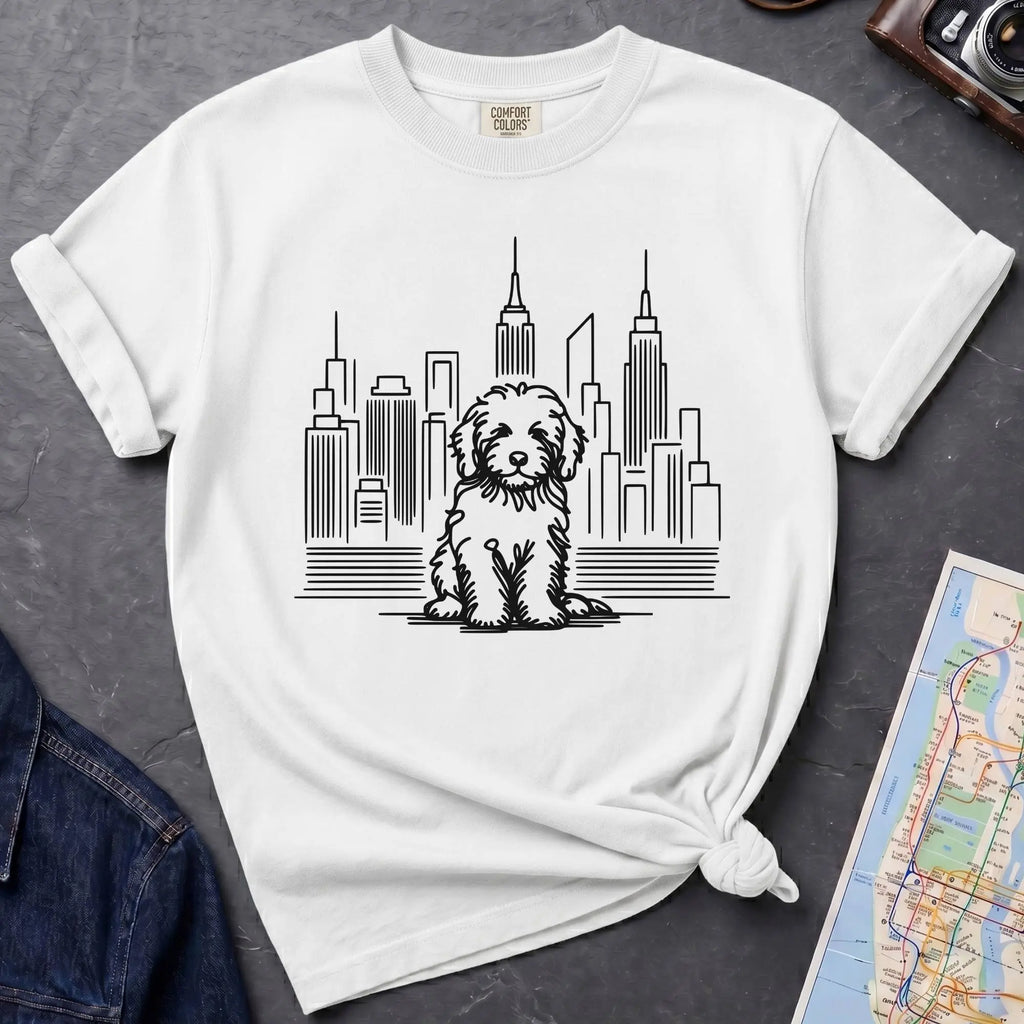 White t-shirt with a Golden Doodle dog and cityscape design on a gray surface with a map and camera.