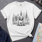 White t-shirt with a Golden Doodle dog and cityscape design on a gray surface with a map and camera.