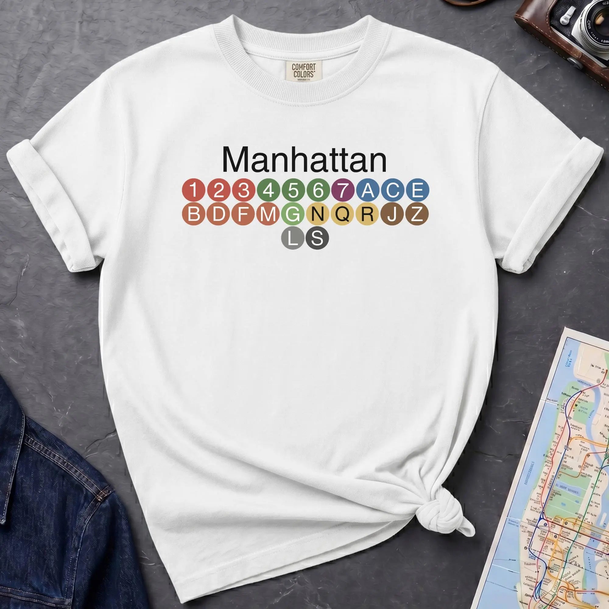 White Comfort Colors t-shirt featuring a minimalist Manhattan subway line graphic with colorful NYC train icons.