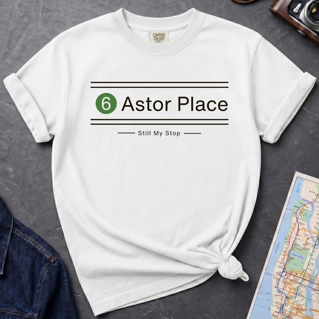 White t-shirt with '6 Astor Place Still My Stop' design on a dark surface with a map and jacket.