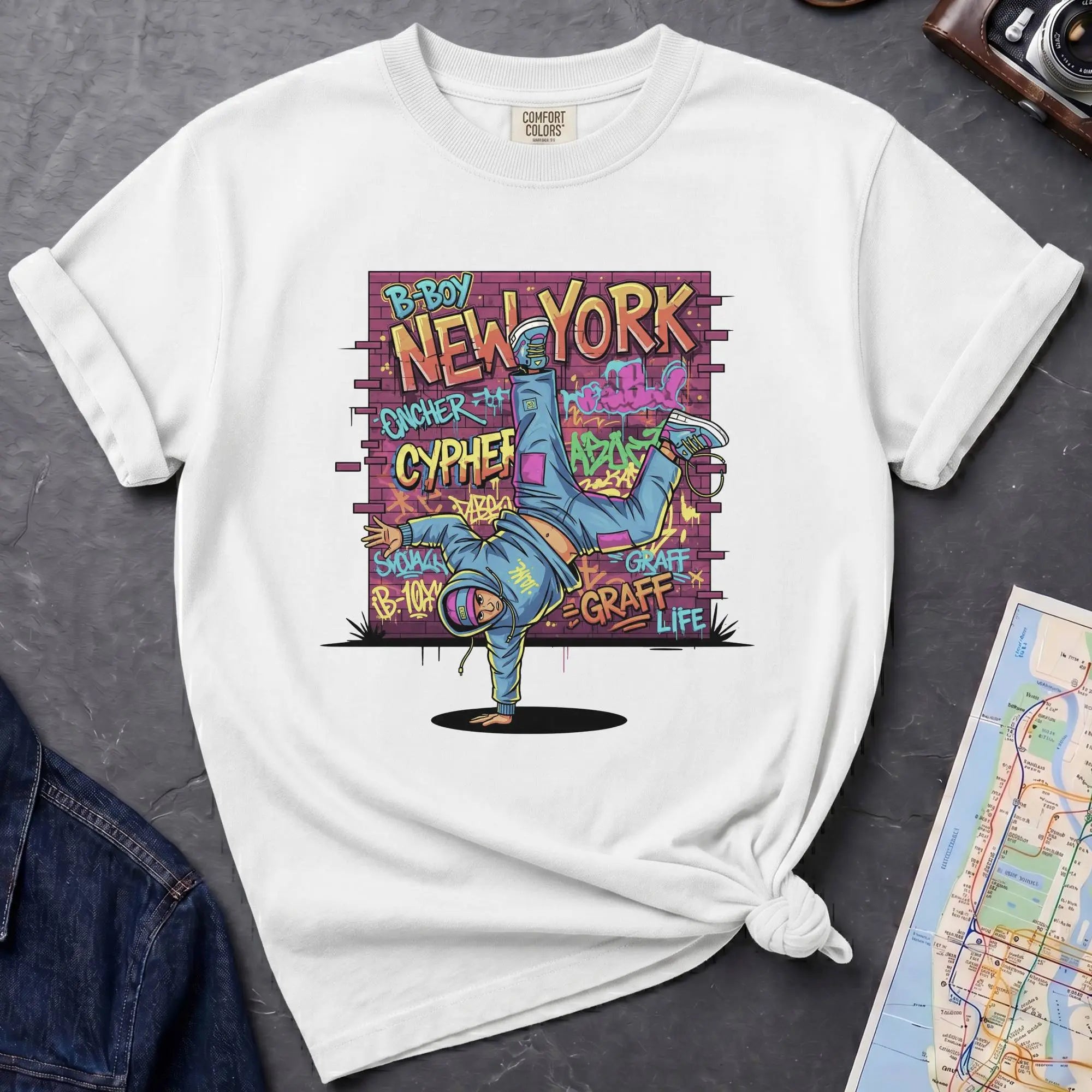 White t-shirt with a colorful graphic design featuring a breakdancer and text on a dark surface with a denim jacket and a map.