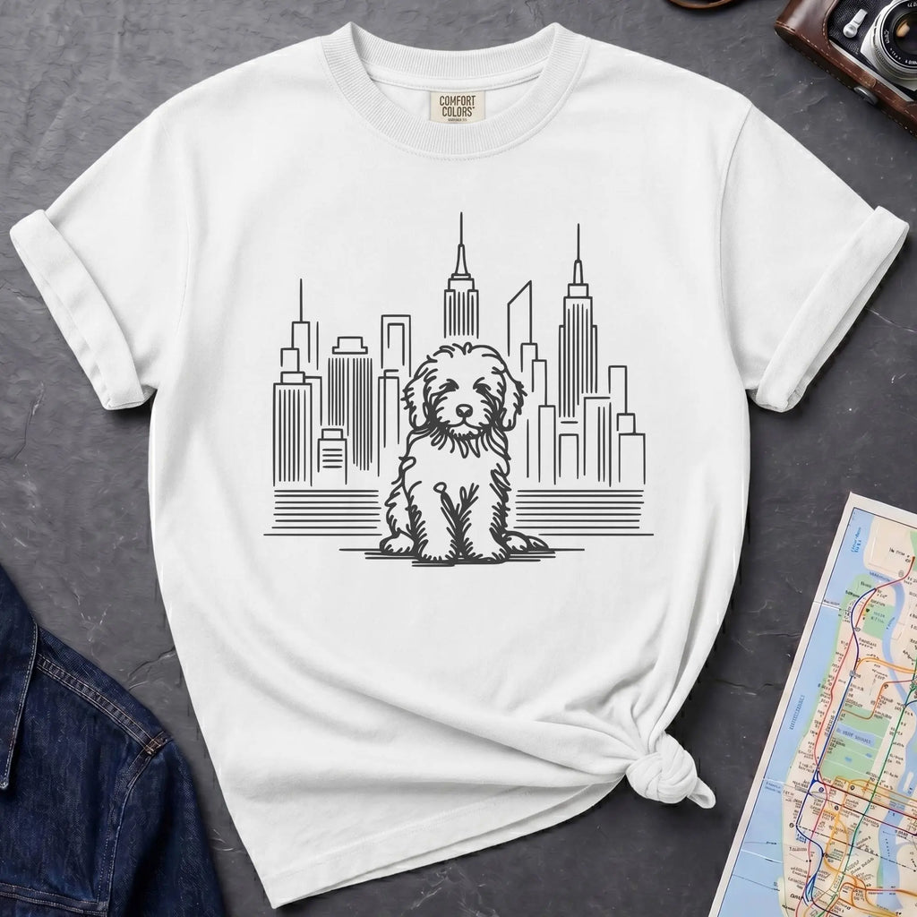 White t-shirt with a Golden Doodle dog and cityscape design on a gray surface with a map and camera.