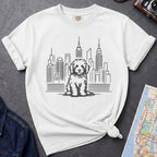 White t-shirt with a Golden Doodle dog and cityscape design on a gray surface with a map and camera.