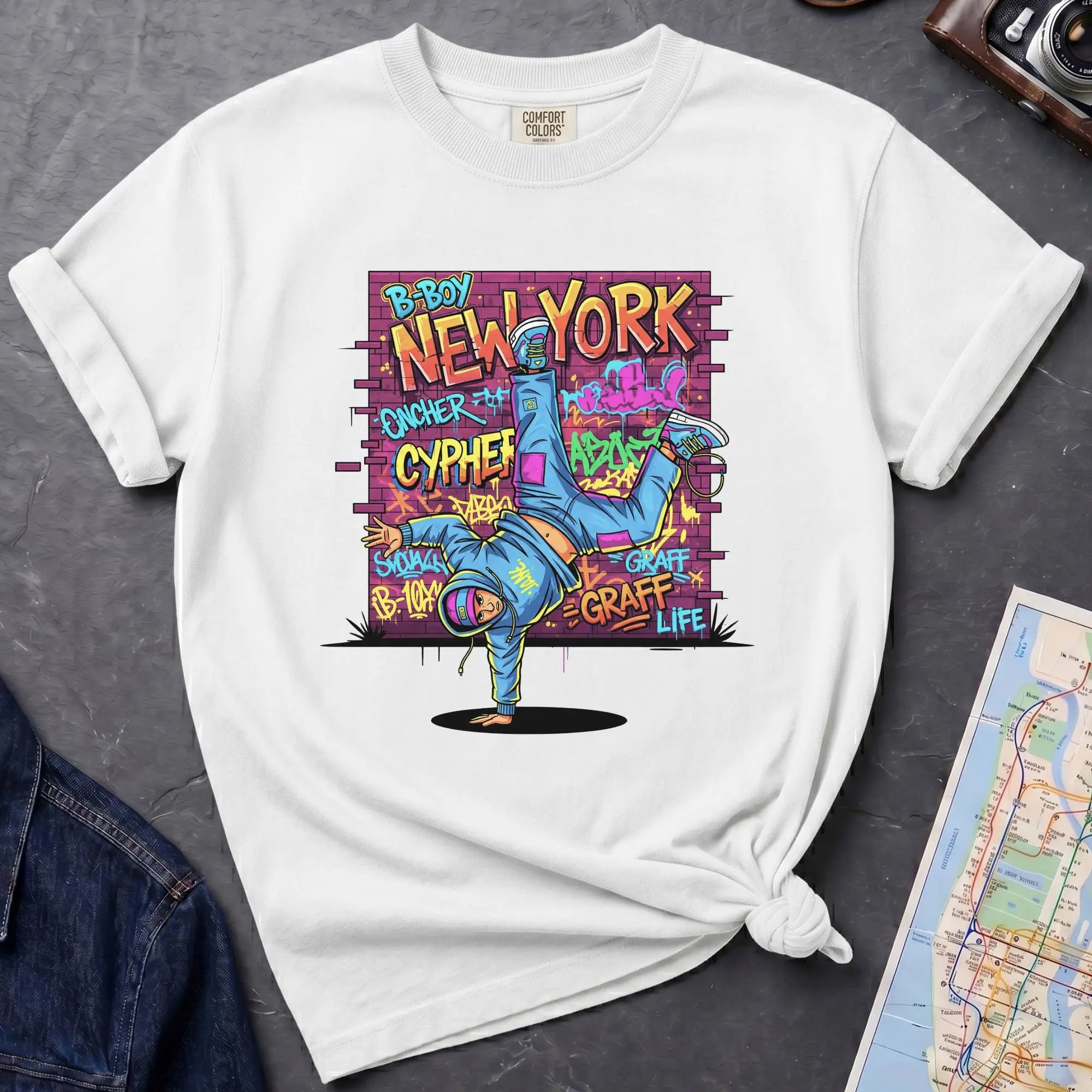 White t-shirt with a colorful graphic design featuring a breakdancer and text on a dark surface with a denim jacket and a map.