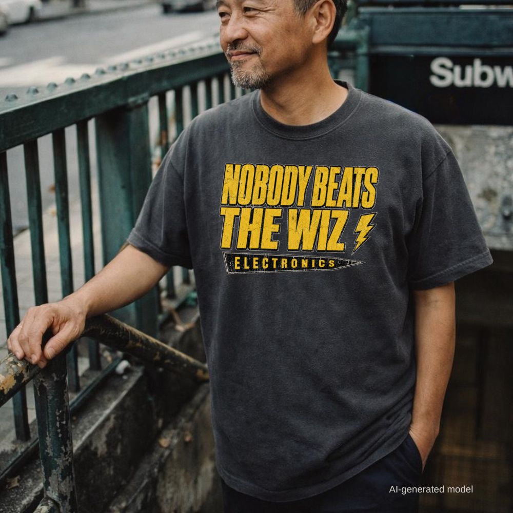 A man wearing a pepper-colored Comfort Colors t-shirt with a bold yellow graphic that reads 'NOBODY BEATS THE WIZ ELECTRONICS' and a lightning bolt, standing outdoors near a subway entrance.