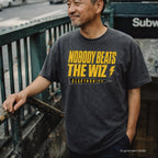 A man wearing a pepper-colored Comfort Colors t-shirt with a bold yellow graphic that reads 'NOBODY BEATS THE WIZ ELECTRONICS' and a lightning bolt, standing outdoors near a subway entrance.