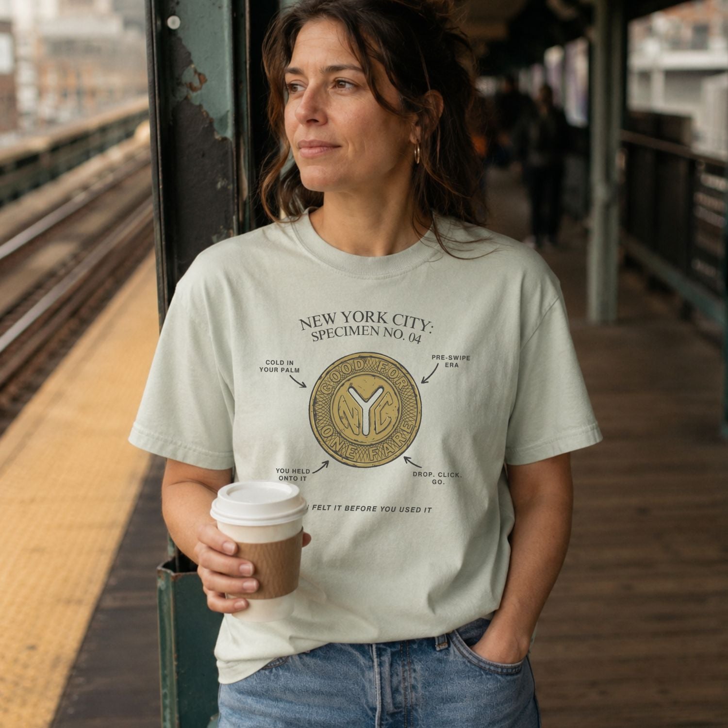 Woman holding a coffee cup on a train platform wearing a Bay colored Comfort Colors t-shirt featuring a vintage New York City subway token graphic with specimen-style callouts and “You felt it before you used it” text.